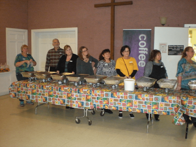 A group of people stand behind food warmers at a table.