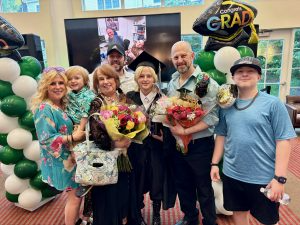 A group of people celebrate a NewBridge JobPlus commencement, holding flowers, balloons, and wearing graduation attire.