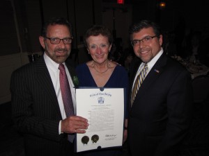 Three people stand together, two men in suits flanking a woman holding an award certificate.