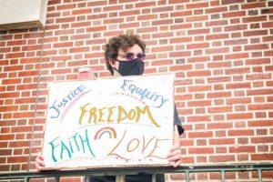 A person in a mask holds a sign against a brick wall. 