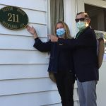 Two masked people stand outside a house, pointing to a sign that reads "PARKER HOUSE 211 MAPLE AVE".