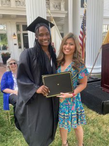 Two women stand together, one in graduation attire holding a diploma