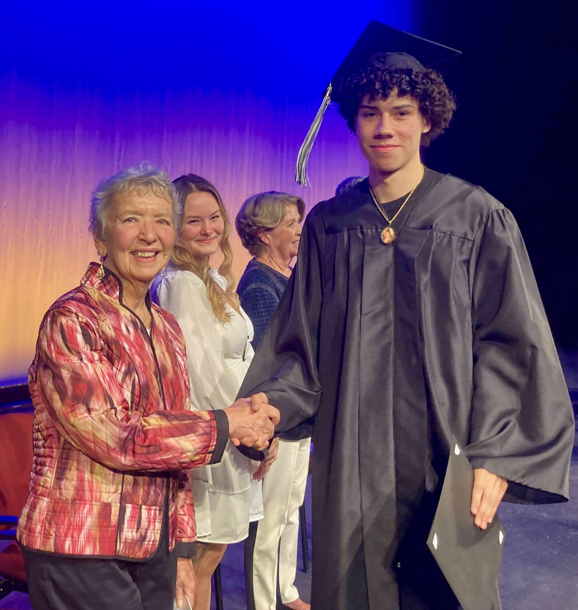 A student in a graduation gown shakes hands with a woman, while other people stand in the background on a stage. 