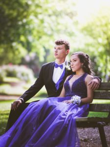 A prom couple sits together on a bench, dressed in a black suit and purple dress.